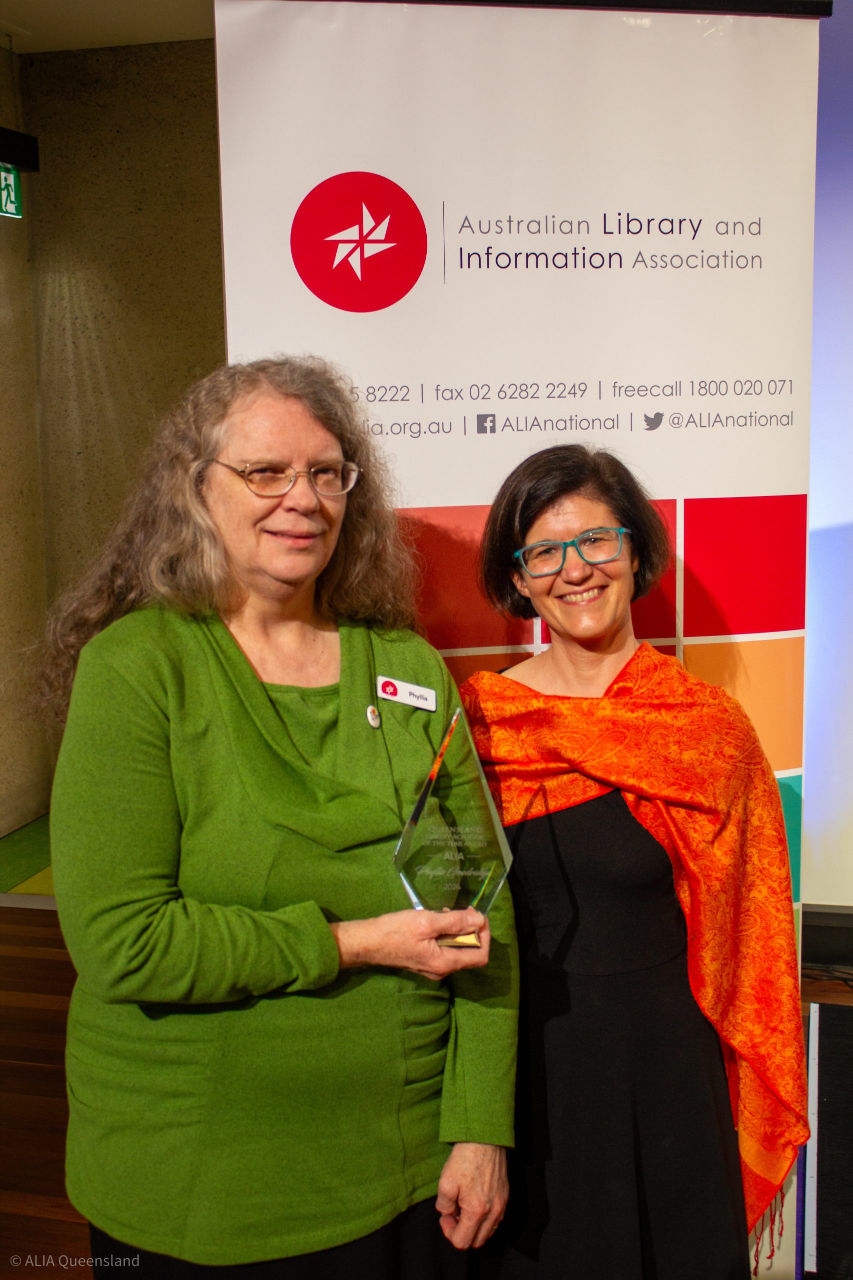 ALIA mincon 4.10.24-002 A woman with fair wavy shoulder length hair wearing a long sleeved light green top stands beside a woman with dark short hair wearing an orange shawl over a black outfit. The woman in green holds a glass trophy. They are standing in front of an ALIA banner.